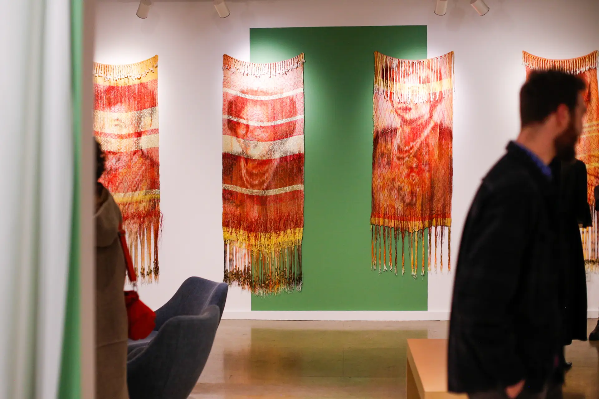 Scene from a gallery with people walking past large, woven art pieces depicting the faces of women.